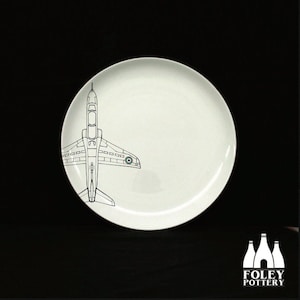 May include: A white ceramic plate featuring a black line drawing of a jet plane. The plane has a roundel on the wing. The plate is made by Foley Pottery.