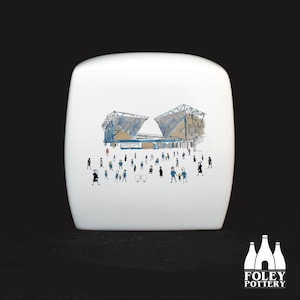 May include: White ceramic storage jar featuring a hand-drawn illustration of a stadium with people in front of it. The stadium is blue and grey with a white roof. The people are all wearing blue and white clothing. The jar has the words "The City" written on it.