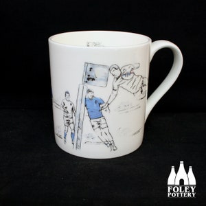 Puede incluir: Taza de cerámica blanca con una ilustración en blanco y negro de un partido de rugby. La ilustración muestra jugadores en acción, con una bandera y un marcador en el fondo.
