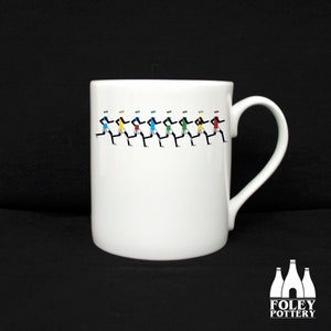 May include: White ceramic mug featuring a black and white design of running figures in colourful jerseys. The mug has a black handle and the text "FOLEY POTTERY" is printed on the bottom.
