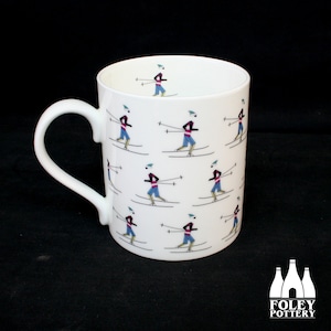 以下が含まれることがあります： 青いズボン、ピンクのジャケット、緑の帽子をかぶったスキーヤーの繰り返し模様が施された白いセラミックマグ。マグには白いハンドルと黒い背景があります。Foley Potteryのロゴが見えます。