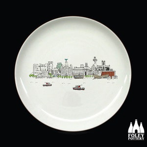 May include: White ceramic plate with a black and white line drawing of the Liverpool skyline. The skyline includes the Liver Building, the Royal Liver Building, and the Anglican Cathedral. There are small boats in the water below the skyline.