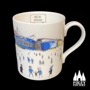 May include: White ceramic mug with a blue and yellow illustration of the Ricoh Arena, a stadium in Coventry, England. The mug has a white handle and the words "Ricoh Arena" are printed on the side. The mug is made by Foley Pottery.