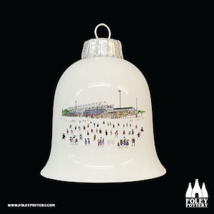 Peut inclure: Ornement de Noël en céramique blanche en forme de cloche avec une illustration en noir et blanc d'un stade avec des personnes devant. L'ornement a un sommet argenté.