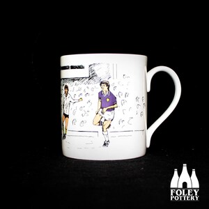 May include: White ceramic mug with a black and white illustration of two soccer players on a field. The mug has a black handle.