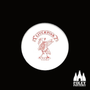 Puede incluir: Posavasos de cerámica blanca con un escudo rojo de Liverpool que representa un pájaro con una rama de follaje en el pico. El texto "LIVERPOOL" está escrito en una pancarta sobre el pájaro. El texto "Liver Bird" está escrito debajo del pájaro.