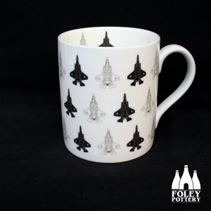 May include: White ceramic mug with a repeating pattern of black and gray jet fighter illustrations. The mug has a curved handle and a glossy finish. The Foley Pottery logo is visible at the bottom right.