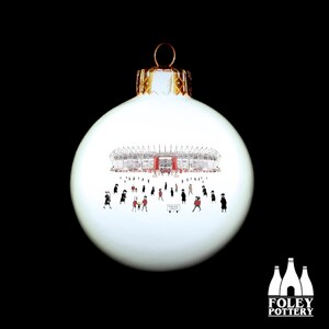 May include: White Christmas ornament with a black and white illustration of a stadium with people in front of it. The stadium is red and white. The ornament has a gold top.