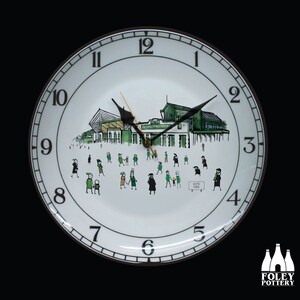 Puede incluir: Un reloj de cerámica blanca con números y manecillas negros. La esfera del reloj presenta una ilustración verde y blanca de un estadio con personas caminando hacia él. El texto "HOME PARK" es visible en un cartel en la ilustración. El reloj está rodeado por un borde marrón.