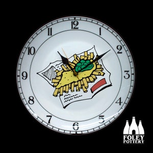 May include: A round wall clock with a white face featuring a whimsical illustration of fish and chips. The clock hands are black, and the numbers are in a classic font. The clock has a black frame.