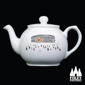May include: White ceramic teapot featuring a black and white illustration of a stadium and people walking around it. The stadium has a red and white crest on the side. The text "Emirates Stadium" is visible on the teapot.