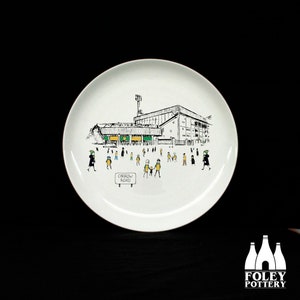 May include: A white ceramic plate with a black and white illustration of a stadium with people walking towards it. The stadium has green and yellow accents. The text "Carrow Road" is visible on a sign in the image.