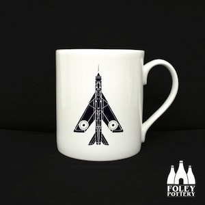 Könnte beinhalten: Eine weiße Keramiktasse mit einer schwarzen Silhouette eines Kampfjets, die auf der Vorderseite aufgedruckt ist. Die Tasse hat einen schwarzen Henkel und wird von Foley Pottery hergestellt.