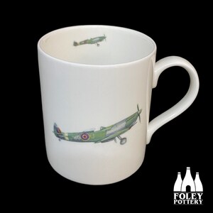 May include: White ceramic mug with a green and brown airplane illustration. The airplane has a red, white, and blue roundel on the side. The mug has a white handle.