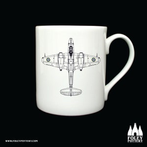 Könnte beinhalten: Weiße Keramiktasse mit einer Schwarzweiß-Zeichnung eines Vintage-Flugzeugs. Das Flugzeug ist nach unten gerichtet, die Flügel sind ausgebreitet. Das Flugzeug hat auf jedem Flügel eine Rundel.