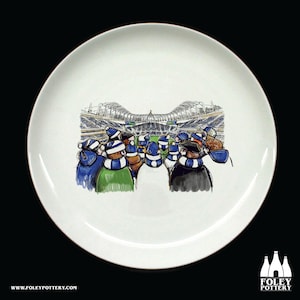May include: White ceramic plate with a hand-drawn illustration of a crowd of people wearing blue and white scarves, looking up at a stadium. The stadium is in the background and is mostly white with some blue details.