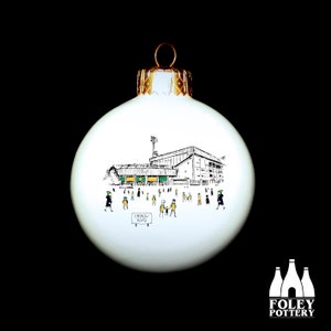 May include: White Christmas ornament with a black and white illustration of a stadium with people in the foreground. The text "Carrow Road" is on a sign in the foreground.