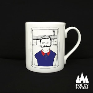 May include: White ceramic mug with a black and white illustration of a man with a mustache wearing a blue shirt. The illustration is framed in black and set against a white background. The mug has a black handle.