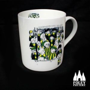 May include: White ceramic mug with a black and white illustration of a crowd of people wearing green and yellow scarves. The mug has a black handle and the text "FOLEY POTTERY" is printed on the bottom.