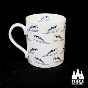 May include: White ceramic mug with a repeating pattern of skiers in mid-air, wearing blue and pink ski suits. The mug has a white handle.