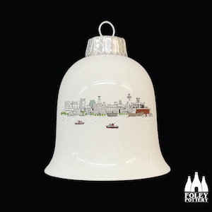 May include: White ceramic Christmas ornament shaped like a bell with a black and white illustration of the Liverpool skyline. The ornament has a silver top.