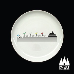 May include: White ceramic plate with a black and white design of cyclists riding up a mountain. The cyclists are in a line and are wearing different colored jerseys. The plate has a black border.