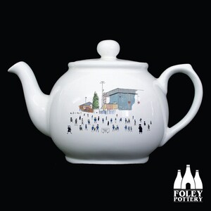 May include: White ceramic teapot featuring a black and white illustration of a stadium and people in blue and black clothing. The text "Stanley Park" is visible on a sign in the illustration.