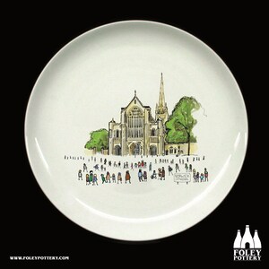 May include: A white ceramic plate with a hand-drawn illustration of Norwich Cathedral in England. The illustration shows the cathedral with a crowd of people in front of it. The text "Norwich Cathedral" is written on a sign in the bottom right corner of the plate.