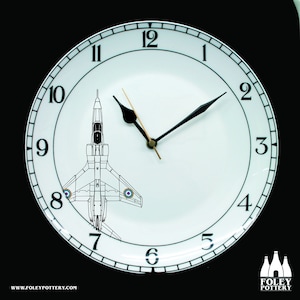 May include: A white ceramic clock plate with black numbers and hands. The clock face features a black and white illustration of a jet plane. The clock is on a black background. The text "WWW.FOLEYPOTTERY.COM" is at the bottom of the plate. The text "FOLEY POTTERY" is at the bottom right of the image.