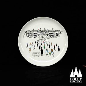 May include: A white ceramic plate with a black and white illustration of Wembley Stadium and a crowd of people walking towards the stadium. The text "Wembley Stadium" is written below the illustration.