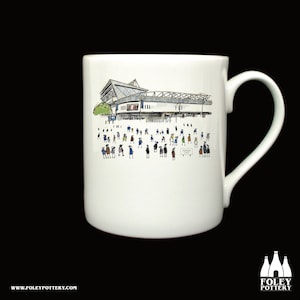 May include: White ceramic mug featuring a black and white illustration of a stadium with people in front of it. The mug has a black handle.