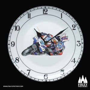 Puede incluir: Un plato de reloj de cerámica blanca con números y manecillas negros. La esfera del reloj presenta una ilustración colorida de un piloto de moto en acción. El reloj está enmarcado por un borde negro con un borde blanco.