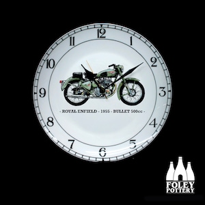 May include: A white ceramic clock face with black numbers and a green Royal Enfield motorcycle illustration. The clock face reads "Royal Enfield - 1955 - Bullet 500cc -" and is surrounded by a black rim with white numbers.