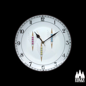 Może przedstawiać: Biały ceramiczny zegar z czarnymi cyframi i wskazówkami. Tarcza zegara przedstawia trzy stylizowane drużyny wioślarskie z kolorowymi wiosłami.