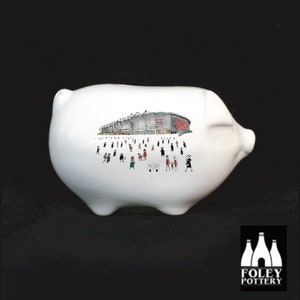 May include: White ceramic piggy bank with a black and white illustration of a stadium with people in the stands. The stadium is red and white. The piggy bank is made by Foley Pottery.