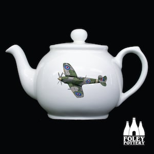May include: White ceramic teapot with a green and brown airplane design. The airplane has red, white, and blue markings. The teapot is made by Foley Pottery.
