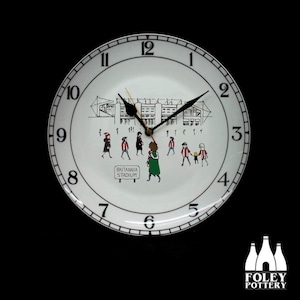 Puede incluir: Reloj de cerámica blanca con una esfera y agujas negras. La esfera del reloj presenta una ilustración en blanco y negro de un estadio con personas caminando hacia él. El texto "BRITANNIA STADIUM" está impreso en la esfera del reloj.