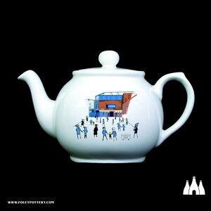 May include: White ceramic teapot with a blue and red illustration of a stadium and people walking towards it. The text "Goodison Park" is printed on the teapot.