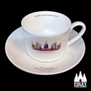 May include: White teacup and saucer set. The teacup features a colorful illustration of a ceremony and the text "May Flights of Angels sing thee to thy rest." The saucer has the text "Her Majesty Queen Elizabeth II 1926-2022."