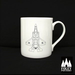 May include: A white ceramic mug with a black line drawing of a fighter jet on the front. The jet is facing down with its wings spread out. The mug has a black handle.