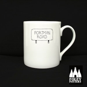 May include: White ceramic mug with a black line drawing of a street sign that says "Portman Road".