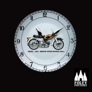 May include: A white ceramic clock with black numbers and hands. The clock face features a black and yellow illustration of a 1964 Ariel Arrow Super Sports 250cc motorcycle. The text "ARIEL - 1964 - ARROW SUPER SPORTS 250cc -" is printed below the motorcycle.