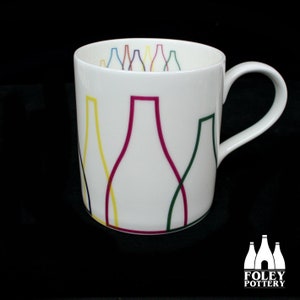 Pode incluir: Caneca de cerâmica branca com um design abstrato colorido de linhas sobrepostas que formam formas de garrafas. A caneca tem uma alça e é feita pela Foley Pottery.