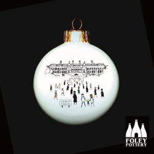 May include: White Christmas ornament with a black and white illustration of Wembley Stadium and people walking towards it. The ornament has a gold top and the text "Wembley Stadium" is printed on the bottom.