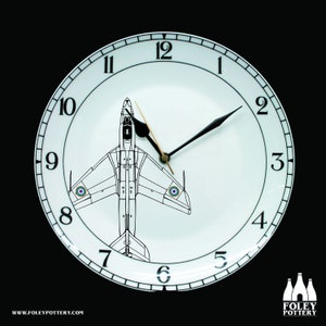 May include: A white ceramic clock plate with black clock hands and a black and white illustration of a jet plane in the center. The clock face has black numbers 1 through 12 around the edge. The text "WWW.FOLEYPOTTERY.COM" is at the bottom of the plate.