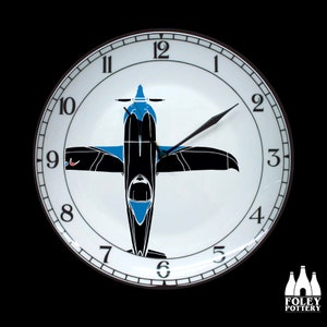 May include: A white ceramic clock with black numbers and hands. The clock face features a black and blue silhouette of a plane. The clock is 12 inches in diameter.