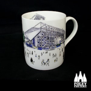 May include: White ceramic mug featuring a detailed illustration of a stadium with a blue and white color scheme. The mug's design includes small figures of people, and the handle is on the right side. The Foley Pottery logo is at the bottom.