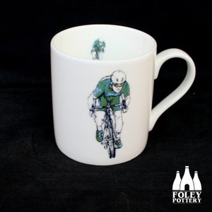 May include: White ceramic mug with a green and blue illustration of a cyclist riding a bicycle. The mug has a white handle and the words "FOLEY POTTERY" are printed on the bottom.