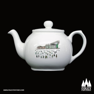 May include: White ceramic teapot with a green and brown illustration of a stadium and people on the side. The teapot has a round body and a spout on the left side. The handle is on the right side.
