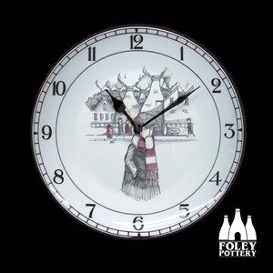 Könnte beinhalten: Eine weiße Keramikuhr mit schwarzem und braunem Rand. Das Zifferblatt der Uhr zeigt eine Schwarzweißillustration einer Person, die eine Straße mit Gebäuden im Hintergrund entlanggeht. Das Zifferblatt der Uhr hat auch schwarze Zahlen und Zeiger. Die Uhr wird von Foley Pottery hergestellt.